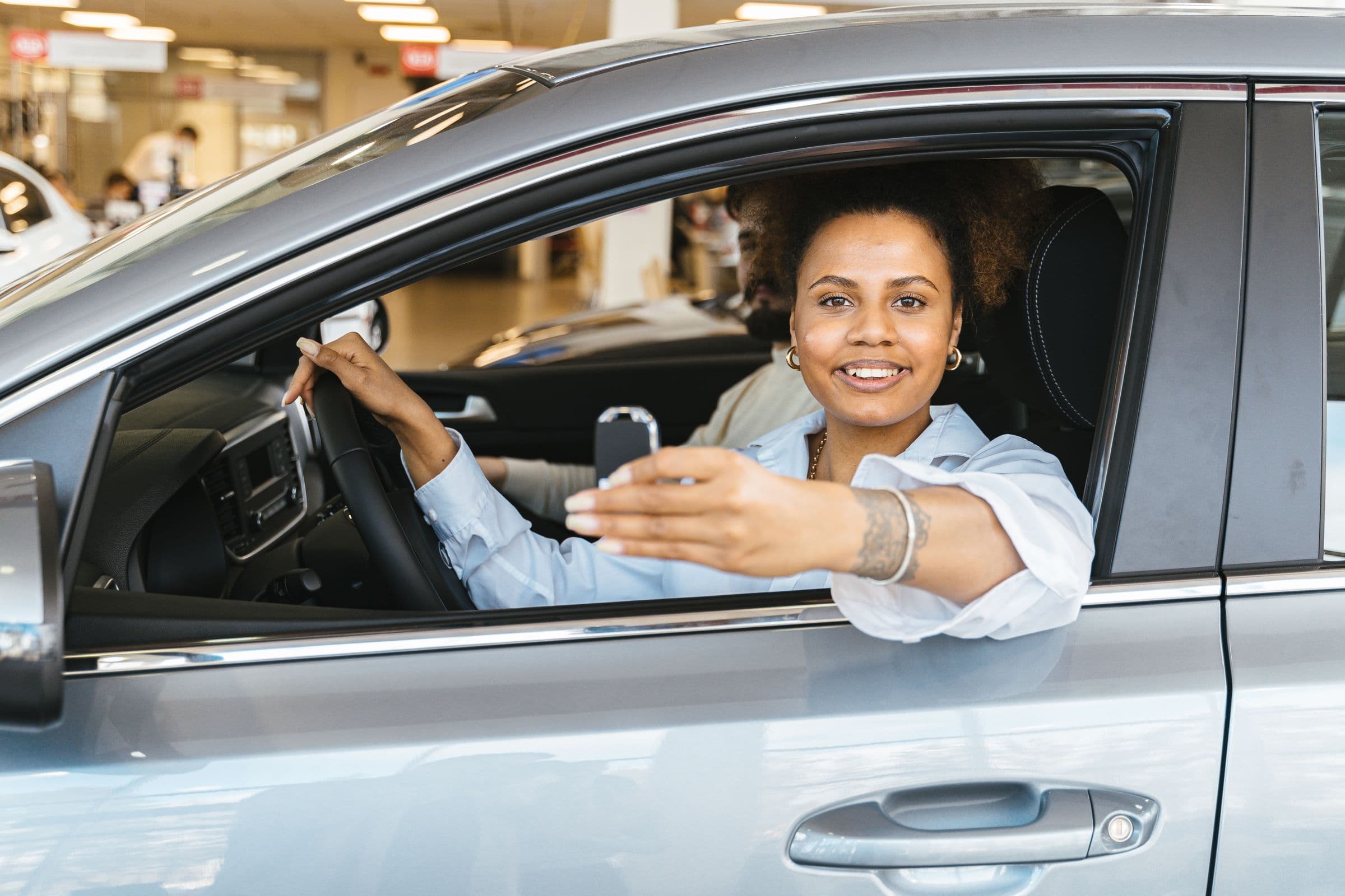 A smiling woman in the driver's seat of a new car at a dealership, holding up the key.